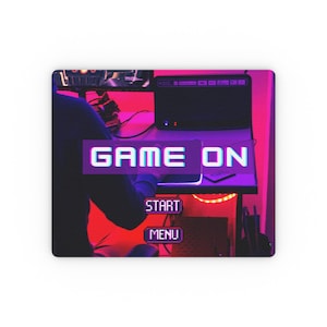 May include: Rectangular mouse mat with a gaming theme. The design features the words "GAME ON" in a pixelated font, along with "START" and "MENU" buttons. The background has a neon pink and purple colour scheme, with a person in the background.