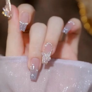 May include: Close-up of a hand with several nails painted with a light pink base and a silver glitter tip. One nail has a small, white, 3D flower design.