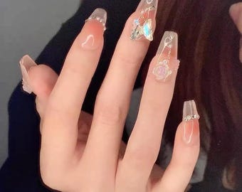 May include: A hand with long, clear acrylic nails. Each nail is adorned with different embellishments, including rhinestones, a butterfly, and a rose design. The nails have a gradient effect, transitioning from a nude color to clear.