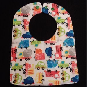 May include: White baby bib with a colorful print of cars, trucks, and construction vehicles. The bib has a green button closure.