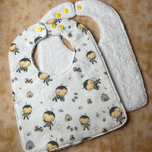 May include: A white baby bib with a pattern of cartoon bees, honeycombs, and leaves. The bib has a terry cloth backing and yellow snap closures. The bib is designed to catch spills and protect clothing.