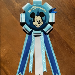 May include: A blue and white rosette featuring a Mickey Mouse button with the text "Walt Disney World." The rosette has multiple layers of blue ribbons, including solid, polka dot, and layered designs. A small blue star accentuates the design.