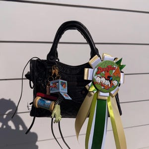 Pode incluir: Bolsa de couro preta com alça superior e fecho de zíper. Um broche DuckTales com fitas verdes, brancas e douradas está preso. Um chaveiro com um personagem de desenho animado está pendurado na alça.