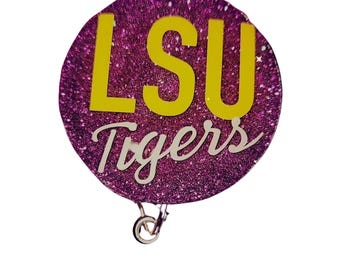 May include: A round, glittery purple badge reel with the text "LSU Tigers" in yellow and white. The badge reel has a retractable cord and a metal clip on the back.