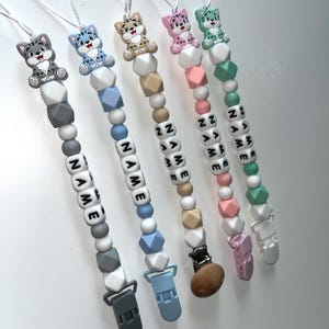 May include: A selection of baby pacifier clips, each with a cat-shaped charm and the name spelled out in letter beads. The clips feature a variety of colours, including grey, blue, beige, pink, and green, with matching clips.