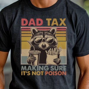 May include: Dark gray t-shirt featuring a raccoon wearing sunglasses, holding a drink and fries. The shirt has the text "DAD TAX MAKING SURE IT'S NOT POISON" in a retro style.