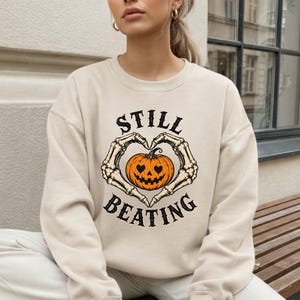 May include: A beige crewneck sweatshirt featuring a graphic print of two skeletal hands forming a heart shape around an orange jack-o'-lantern pumpkin. The text "STILL BEATING" is printed in black capital letters above and below the graphic.