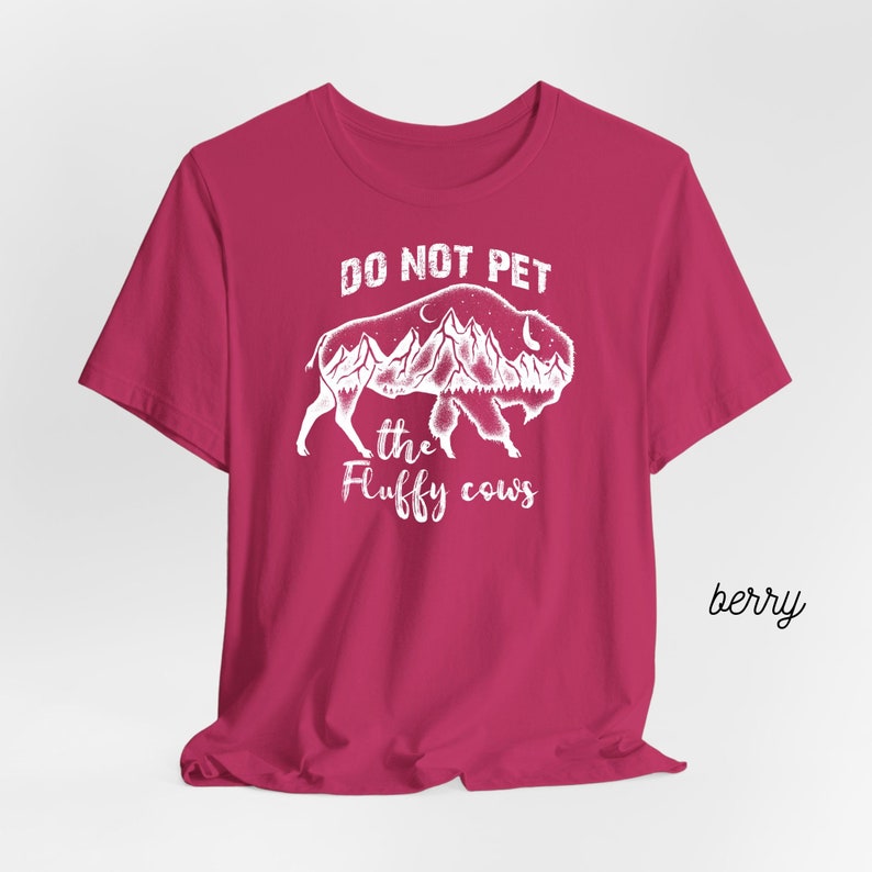 May include: A pink t-shirt with a white graphic of a buffalo with mountains and a moon inside. The text "Do Not Pet the Fluffy Cows" is printed below the buffalo.