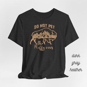 May include: Dark gray heather t-shirt with a brown graphic of a bison with mountains inside its silhouette. The text "Do Not Pet the Fluffy Cows" is printed below the graphic.