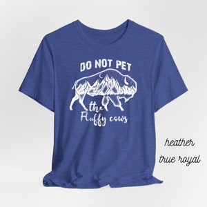 May include: A royal blue t-shirt with a white graphic of a buffalo with mountains and a moon inside its silhouette. The text "Do Not Pet the Fluffy Cows" is printed below the buffalo.