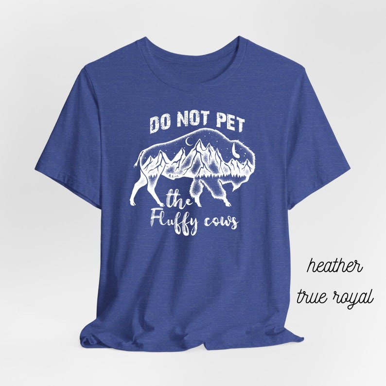 May include: A royal blue t-shirt with a white graphic of a buffalo with mountains and a moon inside its silhouette. The text "Do Not Pet the Fluffy Cows" is printed below the buffalo.