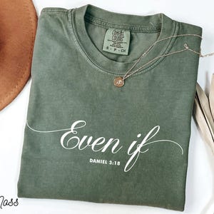 Puede incluir: Una camiseta verde musgo con las palabras "Even if" y el vers&iacute;culo b&iacute;blico Daniel 3:18 en escritura blanca. Un collar dorado con un colgante descansa sobre la camiseta. La camiseta est&aacute; doblada y un sombrero marr&oacute;n es parcialmente visible.