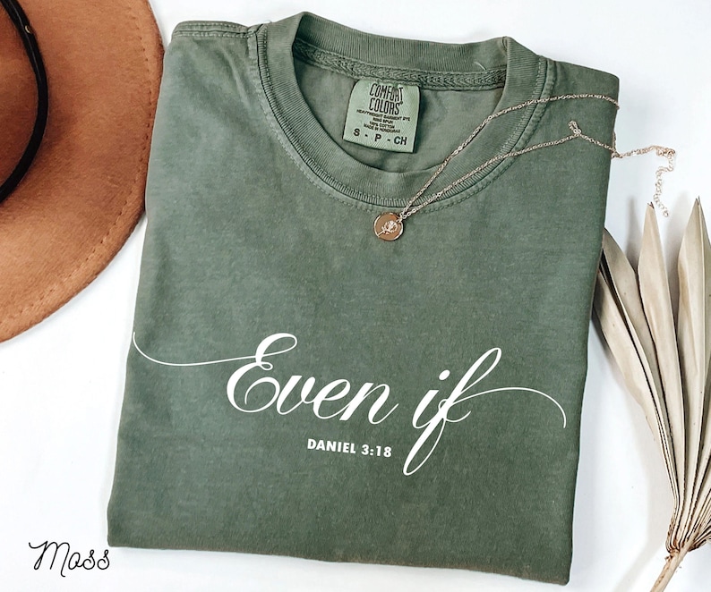 Puede incluir: Una camiseta verde musgo con las palabras "Even if" y el vers&iacute;culo b&iacute;blico Daniel 3:18 en escritura blanca. Un collar dorado con un colgante descansa sobre la camiseta. La camiseta est&aacute; doblada y un sombrero marr&oacute;n es parcialmente visible.