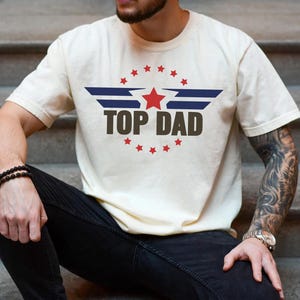 May include: A white t-shirt with a blue and red graphic design featuring a star and wings. The text "TOP DAD" is printed in brown letters.
