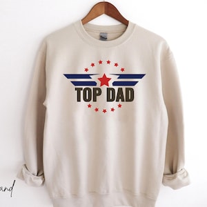May include: A beige sweatshirt with a navy blue, red, and white graphic of a star and wings with the text "TOP DAD".
