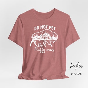 May include: A heather mauve t-shirt with a white graphic of a buffalo with mountains and trees inside its silhouette. The text "DO NOT PET THE FLUFFY COWS" is printed below the buffalo.