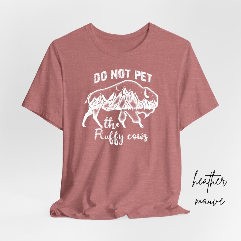 May include: A heather mauve t-shirt with a white graphic of a buffalo with mountains and trees inside its silhouette. The text "DO NOT PET THE FLUFFY COWS" is printed below the buffalo.