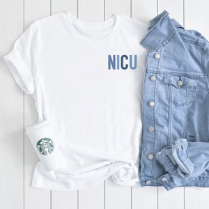 May include: White t-shirt with the word "NICU" printed in blue on the front. The shirt is laid out on a white wooden surface with a denim jacket and a white coffee cup with a green Starbucks logo.