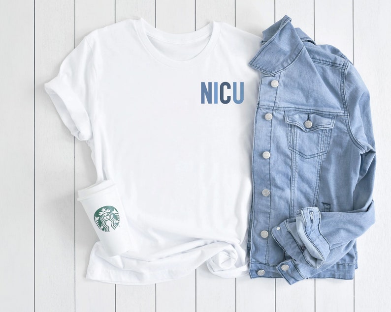 May include: White t-shirt with the word "NICU" printed in blue on the front. The shirt is laid out on a white wooden surface with a denim jacket and a white coffee cup with a green Starbucks logo.