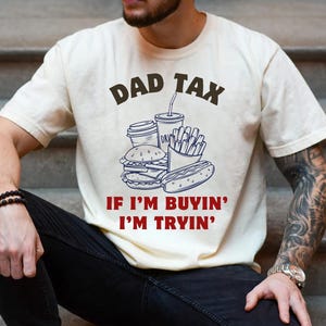 May include: Cream-colored t-shirt with the text "DAD TAX" above a graphic of fast food items, including a burger, fries, and a hot dog. Below the graphic, the shirt reads "IF I'M BUYIN' I'M TRYIN'" in red.