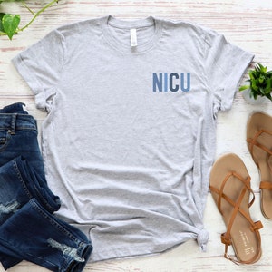 May include: A gray t-shirt with the word "NICU" printed on the front in blue letters. The shirt is laid out on a white wooden surface with a pair of brown sandals and a pair of blue jeans.