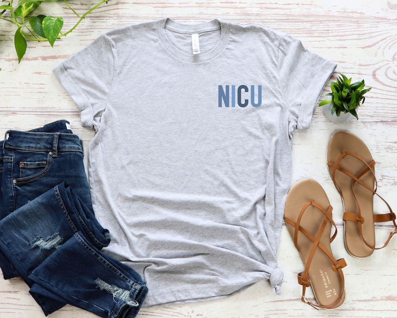 May include: A gray t-shirt with the word "NICU" printed on the front in blue letters. The shirt is laid out on a white wooden surface with a pair of brown sandals and a pair of blue jeans.