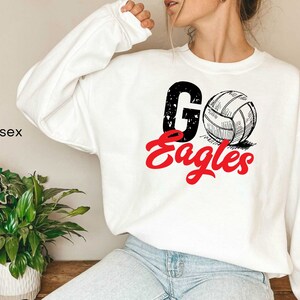 以下が含まれることがあります： 白地に黒と白のバレーボールのグラフィックと赤い文字で「Go Eagles」と書かれたスウェットシャツです。