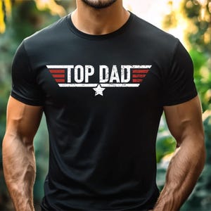 Op de afbeelding: Zwart T-shirt met de tekst "TOP DAD" in het wit, met rode en witte vleugelachtige accenten en een witte ster. Het shirt heeft korte mouwen en is gemaakt van een donker materiaal.