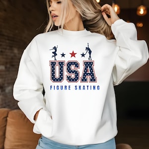 May include: A white long-sleeve crewneck sweatshirt featuring a graphic print with the text USA FIGURE SKATING in blue and red. The design includes silhouettes of two figure skaters, three stars, and the letters USA filled with white stars.