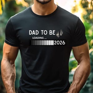 May include: A black short-sleeved t-shirt featuring the white text DAD TO BE, two small baby footprints, the word LOADING, and a progress bar graphic followed by the year 2026 printed across the center of the chest area.