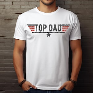 Op de afbeelding: Wit T-shirt met de tekst "TOP DAD" in een verweerde zwarte letter, met rode en zwarte vleugelachtige ontwerpen aan weerszijden. Een zwarte ster is gecentreerd onder de tekst. Het shirt heeft korte mouwen.