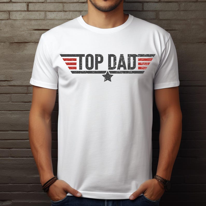 Op de afbeelding: Wit T-shirt met de tekst "TOP DAD" in een verweerde zwarte letter, met rode en zwarte vleugelachtige ontwerpen aan weerszijden. Een zwarte ster is gecentreerd onder de tekst. Het shirt heeft korte mouwen.