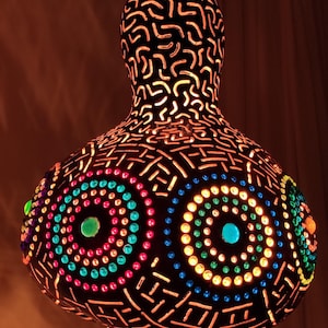 May include: A decorative gourd lamp with a dark brown exterior and intricate cut-out patterns. The lamp features colourful, illuminated circles in red, green, blue, and yellow. The lamp is suspended from a chain.