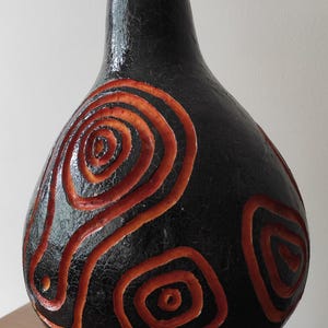 Op de afbeelding: Een decoratieve vaas in de vorm van een kalebas met een zwart, getextureerd oppervlak en oranje spiraalontwerpen. De vaas staat op een houten standaard met een geometrisch patroon. De kalebas heeft een gebogen steel.