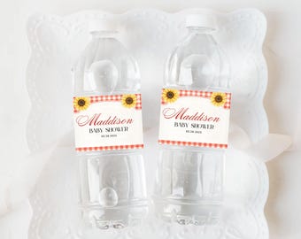 Puede incluir: Dos botellas de agua de plástico transparente con tapas blancas. Cada botella tiene una etiqueta con el nombre "Maddison" en cursiva, "BABY SHOWER" y la fecha 03.28.2025. La etiqueta presenta un patrón de cuadros rojos y blancos e ilustraciones de girasoles.