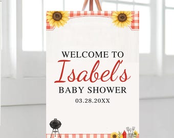 May include: A welcome sign for Isabel's baby shower, featuring a red and white gingham pattern, sunflowers, and barbecue-themed illustrations. The sign includes the text "Welcome to Isabel's Baby Shower" and the date 03.28.20XX.