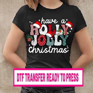 Könnte beinhalten: Schwarzes T-Shirt mit einem farbenfrohen "Have a Holly Jolly Christmas"-Motiv. Das Design zeigt festliche Schriftzüge, Weihnachtsmützen und Lichterketten. Ein rosa Rechteck unten zeigt "DTF TRANSFER READY TO PRESS".