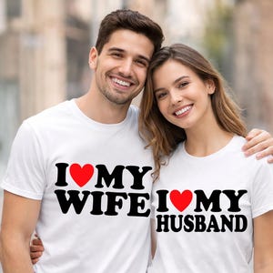 Può includere: Due persone indossano magliette bianche con la scritta "I ❤️ MY WIFE" e "I ❤️ MY HUSBAND" in lettere nere con un cuore rosso. Sorridono e indossano jeans blu.