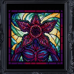Può includere: Un'opera d'arte a punto croce incorniciata raffigurante un Demogorgon della serie Stranger Things. L'opera è realizzata in stile vetrata, con colori vivaci tra cui rosso, viola, verde e giallo. La cornice è nera e ornata.