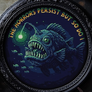 Puede incluir: Un bordado de punto de cruz enmarcado que representa un pez rape pixelado con un señuelo brillante y dientes afilados. El texto "THE HORRORS PERSIST BUT SO DO I!" está bordado alrededor del marco. El marco es negro y ornamentado, sobre un fondo oscuro.