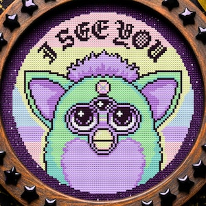 Pode incluir: Uma obra de arte em ponto cruz com uma criatura pixelizada semelhante a um Furby com três olhos, em tons de verde, roxo e rosa. O texto "I SEE YOU" está acima da criatura. Emoldurado em uma moldura redonda de madeira com detalhes de estrelas pretas.