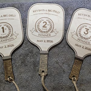 Puede incluir: Tres palas de pickleball de madera grabadas con "KEVIN IS A BIG DILL!" y detalles del torneo. Cada pala muestra un lugar: 1er Campeón, 2do Ganador y 3er Ganador, con fecha del 2 de mayo de 2026. Las palas tienen un acabado de madera clara.