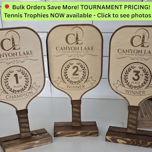 May include: Three wooden tennis trophies with a paddle shape. Each trophy is engraved with "Canyon Lake Junior Women's Club" and a place ranking: 1st, 2nd, and 3rd. The trophies have a dark brown base and handle.