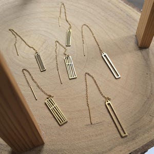 May include: A collection of six gold-toned threader earrings featuring various geometric and leaf-like cut-out designs. Each earring consists of a delicate chain attached to a thin metal bar, with some bars featuring vertical lines or patterns.