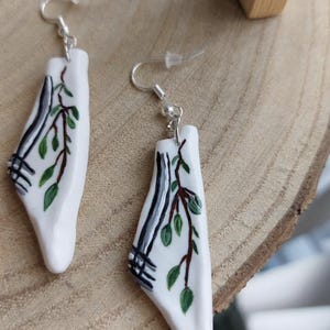 May include: White ceramic earrings with a botanical design. Each earring features a stylized tree branch with green leaves and black and gray accents. The earrings hang from silver-colored hooks.