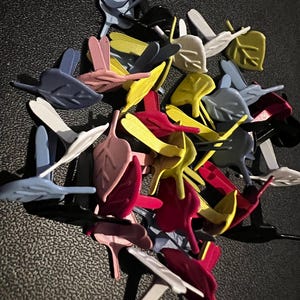 May include: A collection of leaf-shaped plastic clips in various colors, including white, pink, yellow, red, blue, and gray. The clips have a leaf design and a pointed stem. The clips are scattered on a dark surface.
