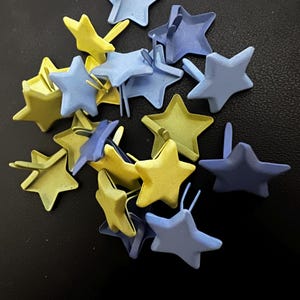 Puede incluir: Un conjunto de chinchetas en forma de estrella en tonos amarillos y azules. Las chinchetas están dispersas sobre una superficie oscura. Cada estrella tiene un pequeño alfiler en la parte posterior. Medidas métricas.