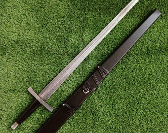 May include: A long, silver sword with a detailed blade and a black handle, resting beside a black leather sheath. The sword has a crossguard and a pointed tip. The sheath has decorative stitching and a buckle.