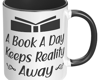 May include: White ceramic mug with a black rim and handle. The mug has a black graphic of an open book and the text "A Book A Day Keeps Reality Away".