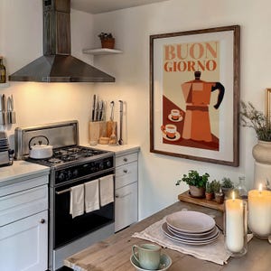 May include: A kitchen interior with a framed poster saying "Buongiorno" and a coffee pot illustration. The kitchen has a stove, cabinets, and a wooden table set with plates and a coffee cup. Two large candles are lit on the table.